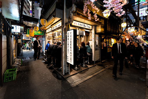 Shinjuku, Japan - April 4, 2019: Memory lane alley with izakaya pub restaurant, business people and businessman sitting and eating by sidewalk in Tokyo city at night