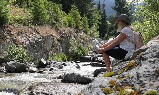 istock Woman with backpack sits on a rock and soaks her feet in a mountain stream 1161861452