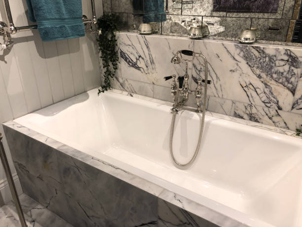 istock Image of luxury bathroom suite with white / grey marble wall and floor tiles, white ceramic bath, sink, WC toilet, washroom with stainless steel Victorian mixer tap and shower plumbing, artificial trailing plants, mirrors, towel rail, tiled floor flooring 1161863234