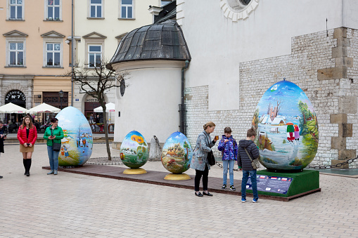 Tourists and locals alike in Krakow, Poland visit an Easter Festival during Holy Week in Rynek Glówny, the 13th century Town Square.  People enjoy beverages, local foods, and homemade crafts.  A family looks at an Easter egg display.