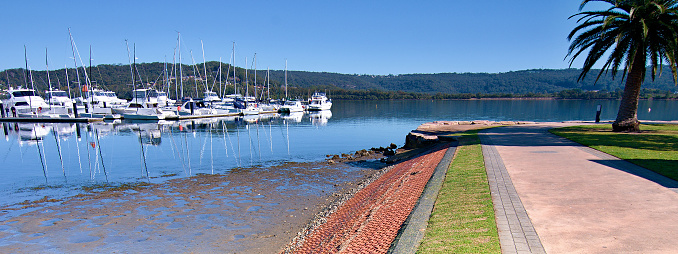 Waterfront park marina/dock panorama landscape with boats, tree covered horizon and a mid blue sky. Safe haven for sailing and cruising vessels. Gosford, Australia.