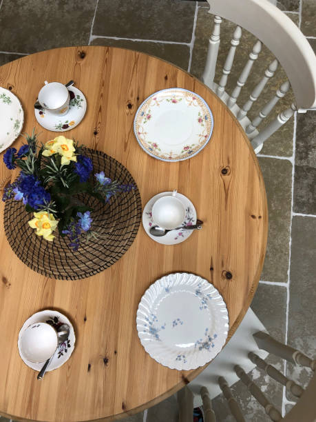 istock Image of circular round pine dining kitchen table with four chairs painted white set for breakfast / lunch with dinner plates, tea cups and saucers, vase of flowers and placemats, on tiled floor with stone tiles, kitchen cabinets / cupboards in background 1161917055