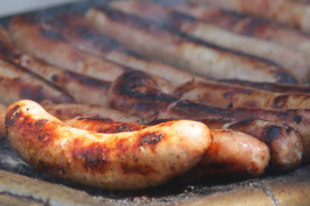 Stock photo of garden party charcoal barbecue large sausages in row cooking / grilling perfectly on griddle grill, burning smoke, smoky BBQ with burger serving tongs turning each sausage, English bangers being cooked for barbecued hot dogs at street market food imagen de la fiesta del jardín de carbón barbacoa salchichas grandes en la fila cocinar / asar perfectamente en la parrilla de la plancha, humo ardiente, barbacoa ahumada con chuseles que giran cada salchicha, bangers ingleses que se cocinan para perros