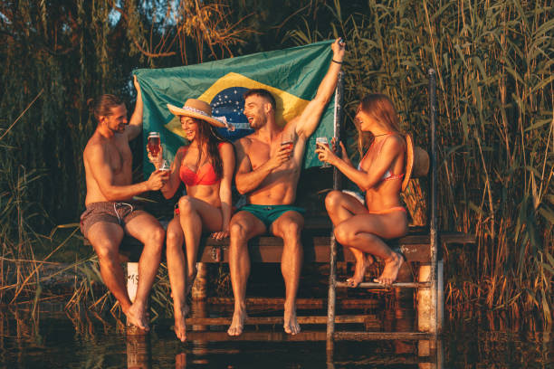 счастливая группа молодых людей, пьющих пиво с бразильским флагом на скамье подсудимых у реки в летний солнечный день - brazil serbia стоковые фото и изображения