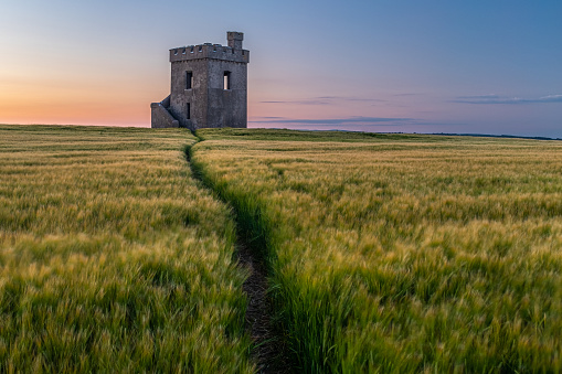 A lookout fort standing proud in a field of wheat at sunset, path in the field leading to the fort nobody in the image