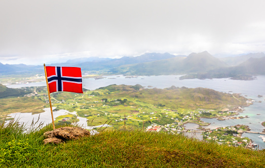 Norwegian national flag in the wind on the top of Nonstinden peak with fjord in the background, Ballstad, Vestvagoy Municipality, Nordland county, Norway
