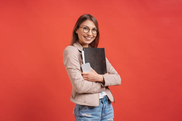 istock Young smiling student or intern in eyeglasses standing with a folder on red background. 1161932422