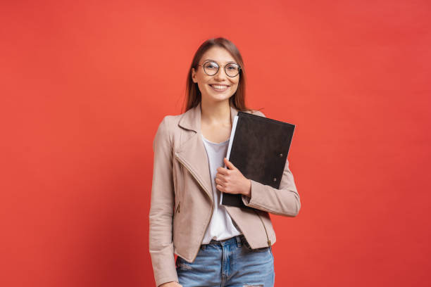 Young smiling student or intern in eyeglasses standing with a folder on red background. stock photo