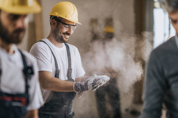 Happy construction worker cleaning his gloves from sawdust at in renovating apartment. Young happy manual worker cleaning sawdust from his protective gloves at construction site. menial job stock pictures, royalty-free photos & images