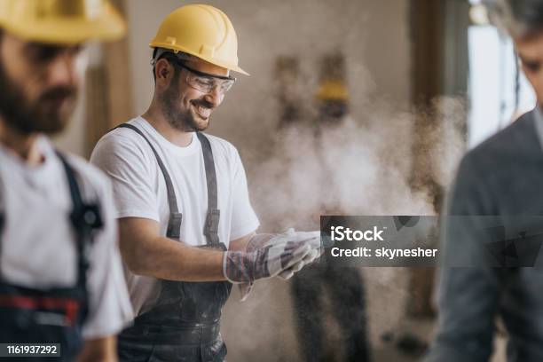 Happy Construction Worker Cleaning His Gloves From Sawdust At In Renovating Apartment Stock Photo - Download Image Now