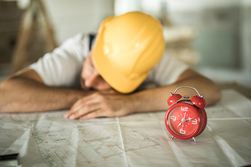 Tired manual worker taking a nap on blueprints at construction site. Focus is on alarm clock.