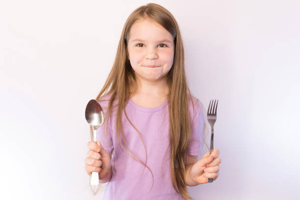istock Little cute girl holding a spoon and fork with a satisfied expression on her face, on a light background 1161956371
