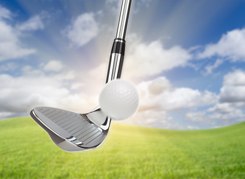 Chrome Golf Club Wedge Iron Hitting Golf Ball Against Grass and Blue Sky Background.