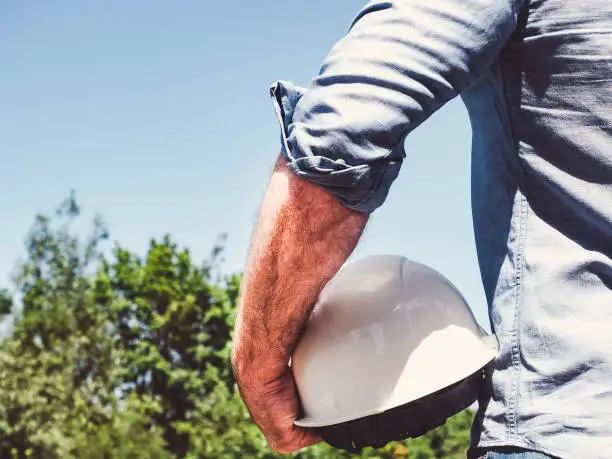 Handsome engineer, holding white hardhat in the park against the backdrop of green trees and the setting sun, looking into the distance. Close-up. Concept of labor and employment