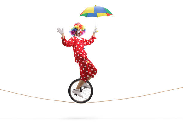 Full length profile shot of a clown riding a unicycle on a rope and holding an umbrella isolated on white background