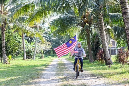 A Malay Muslim girl riding on her bicycle with Malaysian flags attached to it, celebrating Malaysia's National Day.