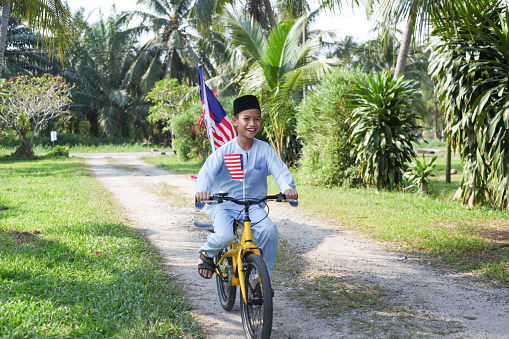 A Malay Muslim boy riding on a bicycle with Malaysian flags attached to it, to celebrate Malaysia's National Day. He is wearing the traditional Malay costume.