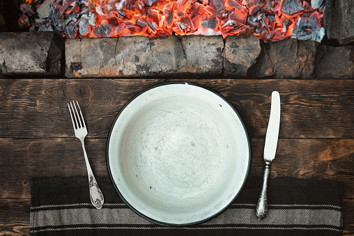 Empty white metal bowl with a copy space for food, towel with a fork and knife on a wooden kitchen table flat lay background with a burning coals above.