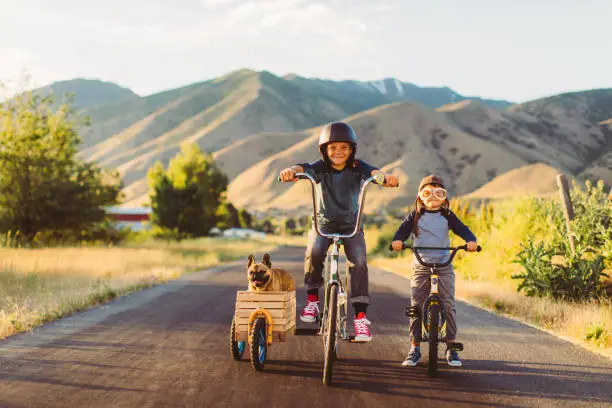 Two young boys with vintage clothing and motorcycle helmets rides their bicycles with a pet and best friend French Bulldog riding along in a side car in Utah, USA. Sometimes a road trip journey with your best friends and some fresh air in your face is the best medicine for the soul.