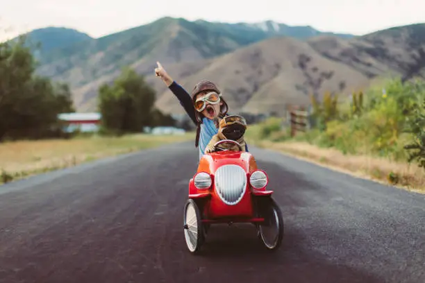 A young boy with flying goggles and flight cap races a red toy car with his pet and best friend French Bulldog along a small road in Utah, USA. Sometimes a road trip journey with your best friend and some fresh air in your face is the best medicine for the soul.