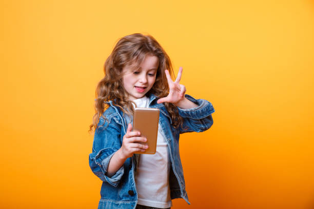 Little girl taking a selfie photo with a smart phone on yellow background. Concept of using modern technology by children. Close-up portrait of cute curly beautiful girl taking a self portrait on yellow background free girl download stock pictures, royalty-free photos & images