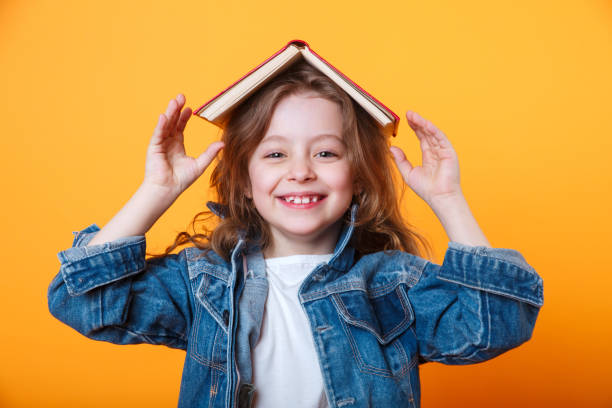 mała uczennica z książką na głowie, ubrana w dżinsową kurtkę i czarne okulary, stojąca na żółtym tle. - child excitement awe fun zdjęcia i obrazy z banku zdjęć