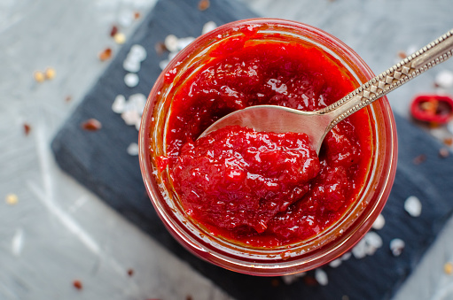 Red hot chili jam in glass jar with fresh ingredients on grey concrete background. Natural homemade peppers sauce with spoon. Fresh Homemade salsa dip. Top view. Close up.