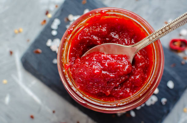 Red hot chili jam with fresh ingredients Red hot chili jam in glass jar with fresh ingredients on grey concrete background. Natural homemade peppers sauce with spoon. Fresh Homemade salsa dip. Top view. Close up. condiment stock pictures, royalty-free photos & images