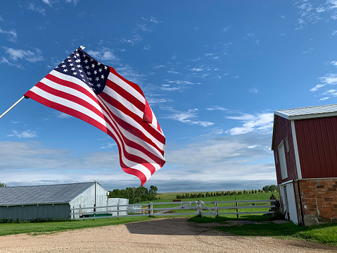 Low angle close up view of American Flag against a blue sky with fluffy white clouds with american farm buildings in background