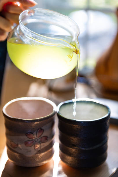 closeup of person hands holding glass teapot pouring japanese sencha green tea in cups or teacups at home for breakfast morning by window - 11315 imagens e fotografias de stock