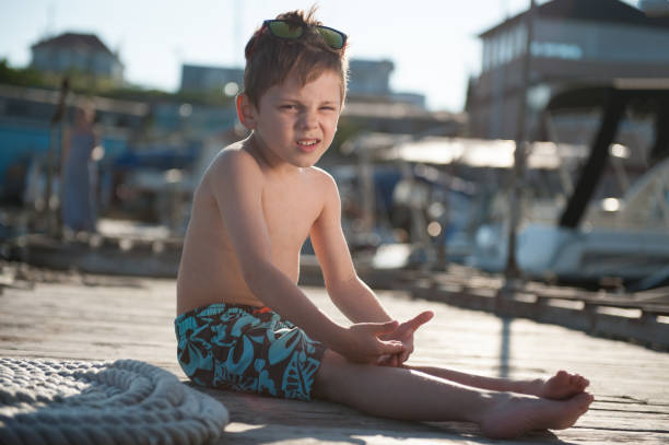 funny healthy cute little adorable boy in shorts sitting on wooden sea pier on sunny summer day squinting - fashionable the human body short hair human head imagens e fotografias de stock