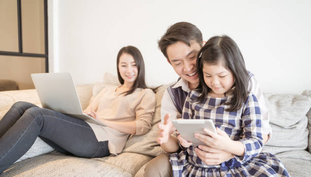happy asian family spending time together on sofa in living room. family and home concept - father digital tablet asian ethnicity daughter imagens e fotografias de stock