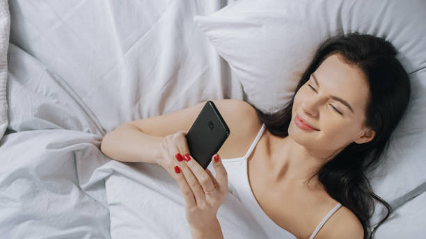 istock Portrait of Beautiful Brunette Using Touchscreen Smartphone in Her Bed and Smiling, Chatting with Friends, Using Social Media, Posting Pictures, Sharing Thoughts. Top Down Shot. 1162082369