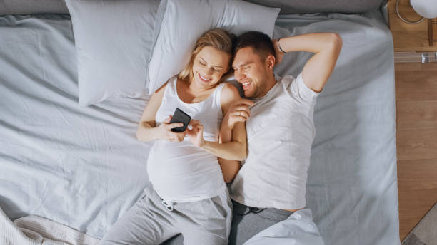 любящая молодая пара, проводя утро в постели, беременная молодая женщина показывает своему партнеру что-то на сенсорном смартфоне. поиск де - проводя поиск стоковые фото и изображения