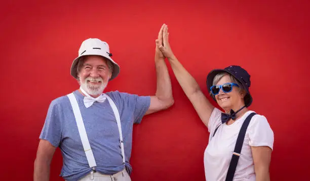 Senior couple of people against a red wall enjoying freedom and holiday. Dressed with colorful caps, bow ties and suspenders. White hair and beard for man