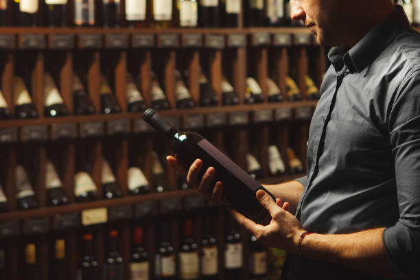 Close up portrait of sommelier holding wine bottle on wine cellar background. Sommelier holding big wine bottle in hands and looking attentively on it, photo in cellar having collection of containers with alcoholic beverages, dim light and handsome man reading label sommelier photos stock pictures, royalty-free photos & images
