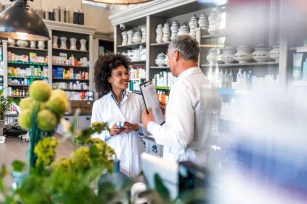 Rear view of mature gray hair male expert with clipboard and female pharmacist after inspection in pharmacy talking to each other with smile. Two smiling friendly male and female pharmacists working in luxury pharmacy. Medicine, healthcare and technology concept - Mature men and mixed race female apothecaries with clipboard and medication at pharmacy.