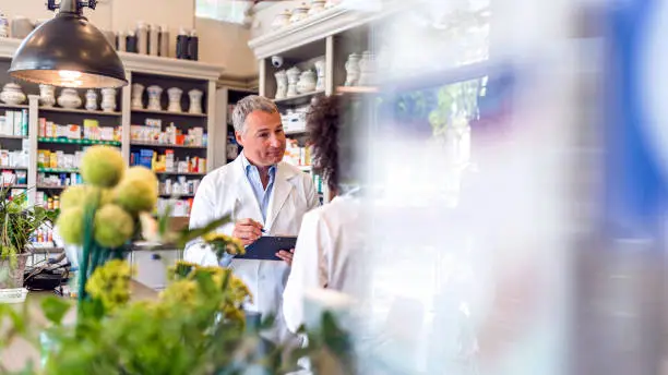 Photo of mature gray hair male expert with clipboard and female pharmacist after inspection in pharmacy talking to each other with smile. Two smiling friendly male and female pharmacists working in luxury pharmacy. Medicine, healthcare and technology concept - Mature men and mixed race female apothecaries with clipboard and medication at pharmacy.