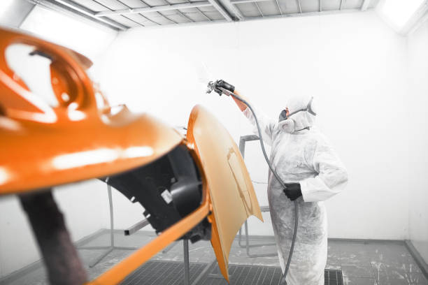 l'uomo con maschera protettiva e vestiti spruzza vernice al cofano dell'auto con una pistola a spruzzo in una cabina di verniciatura. - painting booth foto e immagini stock