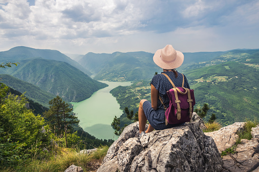 Young traveler girl sitting on mountain cliff looking to stunning view on lake valley.