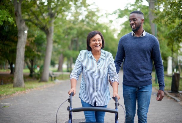 My therapist has helped me walk! Cropped shot of a happy mature woman using a walker while a handsome young man assists her in the park mobility walker stock pictures, royalty-free photos & images