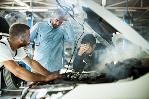 Mid adult customer and mechanics feeling frustrated while the car is smoking in a repair shop.