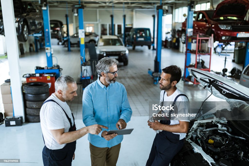 Mid adult manager using touchpad and talking to car mechanics in a workshop. Mid adult manager using digital tablet while talking to mechanics in auto repair shop. Auto Repair Shop Stock Photo