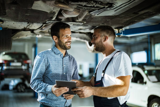 Happy manager and auto mechanic talking while using touchpad in a workshop. Happy male customer and mechanic talking while using digital tablet in auto repair shop. auto repair shop mechanic digital tablet customer stock pictures, royalty-free photos & images