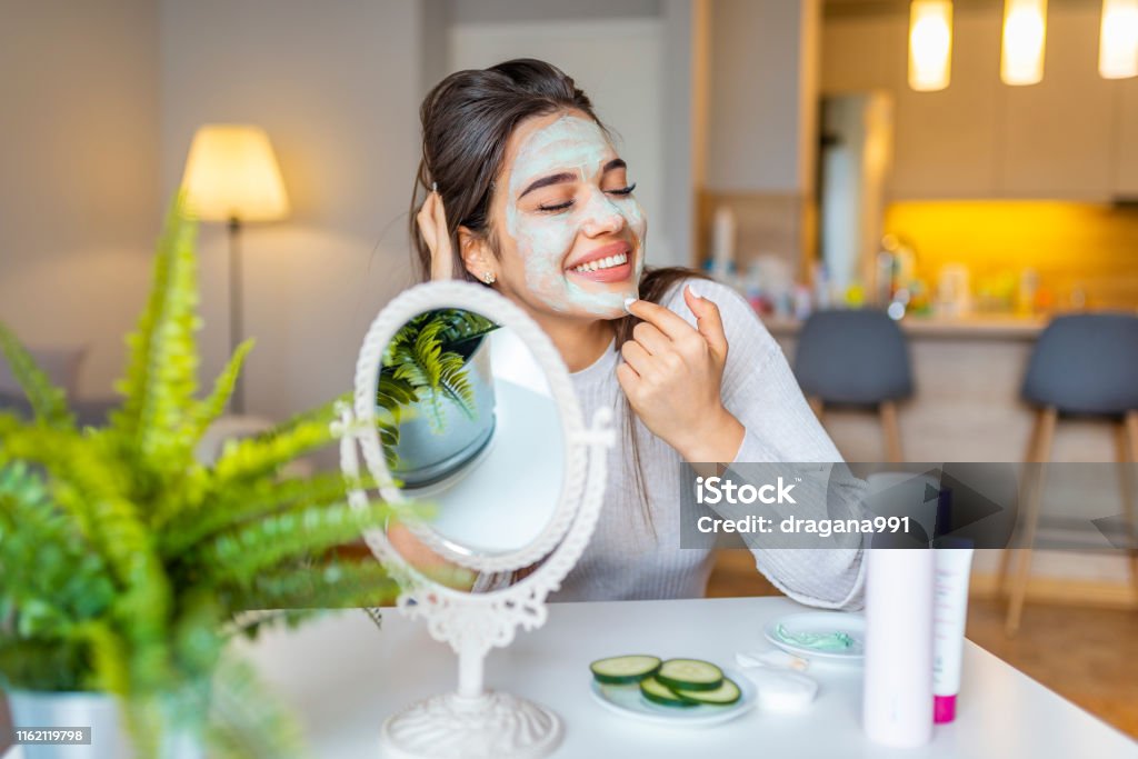 Attractive young woman sitting with a facial mask on her skin Young woman with cleansing mask on her face at home. Skin care. Woman aplying beauty mask,close up. So beautiful. Close-up of girl with beauty mask on her face looking in mirror. Facial Mask - Beauty Product Stock Photo