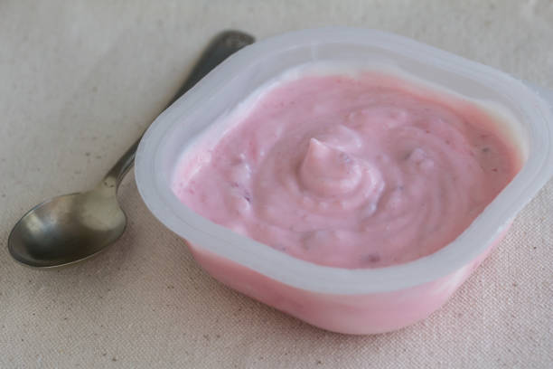 Strawberry yogurt in white plastic cup on linen background with small silver spoon