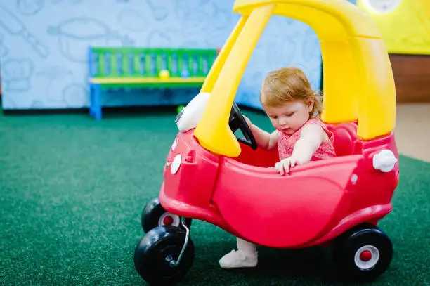 Photo of Happy children, little girl child rides on a red big car on the road. Close up. The baby is driving the car and played in a children's playroom on a birthday party. Time together at entertainment centre.