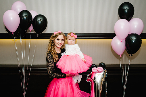 Mother and daughter in a skirt with pink tulle against a background of black and pink balloons. Woman and little baby girl princess. Concept of birthday celebration is 1 year.