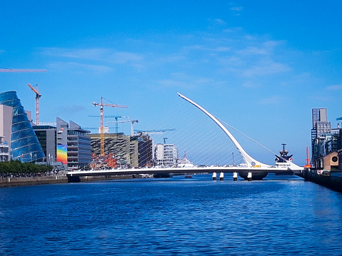 Dublin, Ireland-Juin 26, 2019
Foreground beauty of the dark blue Liffey River in summer contrasting with the whiteness of the Samuel Beckett Bridge, designed by architect Santiago Calatrava and its shape evoking a Celtic harp on the side.
Back side cosmopolitan and dynamic economic Dublin.
With the Convention Center facade in the colors of the LGBTQ Convention on the left, cranes and buildings.
On the right are the Docks and a tourist ferry.
The bridge connects Macken Street to Guild Street and North Wall Quay.