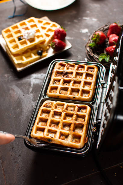 horizontal image of brakfast waffles cooking to a golden brown in a waffle iron with syrup and bowl of fruit sitting on the side..